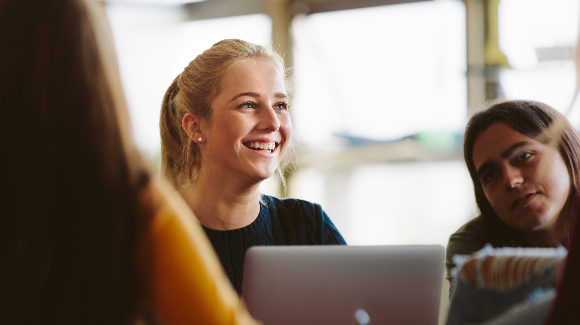 Flicka sitter framför laptop. Hon ler. En person i förgrund och en person i bakgrund. Foto: Istock