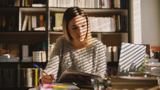 Kvinna vi ett bord. Laptop framför henne. Bokhylla i bakgrunden. Foto Istock