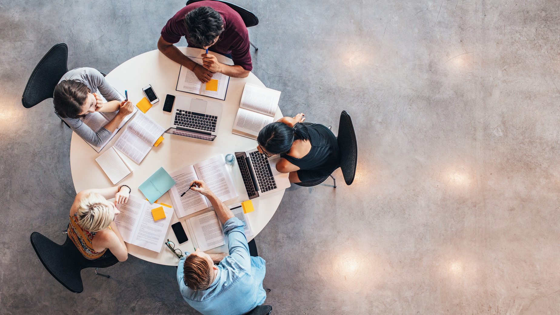 Fem studenter sitter runt ett runt vitt bord. På bordet finns böcker, paper och laptops. Ovanifrånperspektiv. Foto Istock.