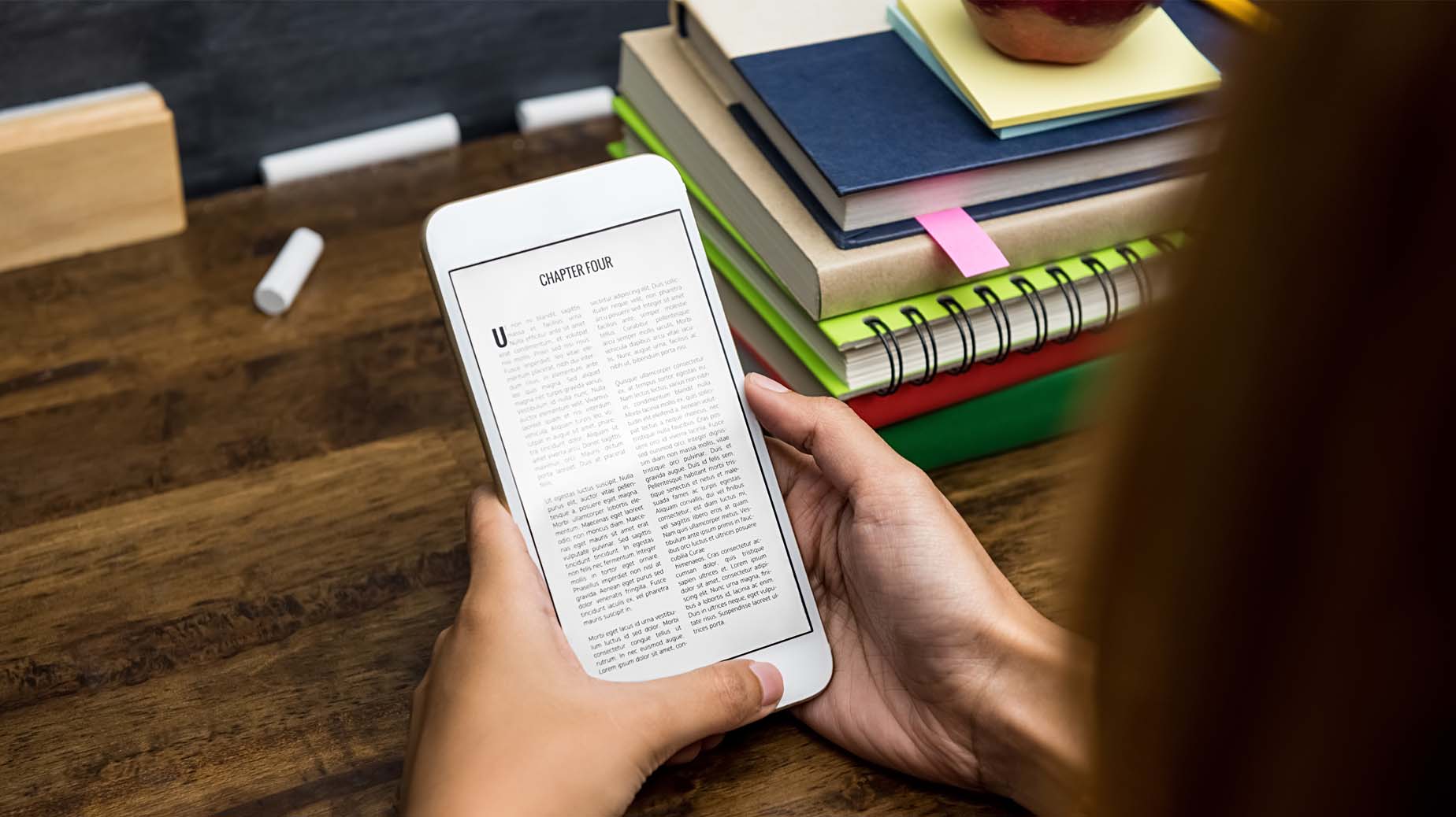Två händer och mobiltelefon med text, i bakgrunden syns böcker och block, foto Istock
